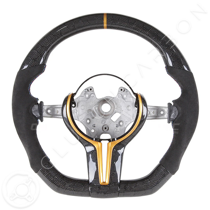 The BMW F-Series Custom Carbon Fiber Steering Wheel is styled with a suede finish, metallic gold center accent, central mounting plate, and an orange stripe atop a white background.